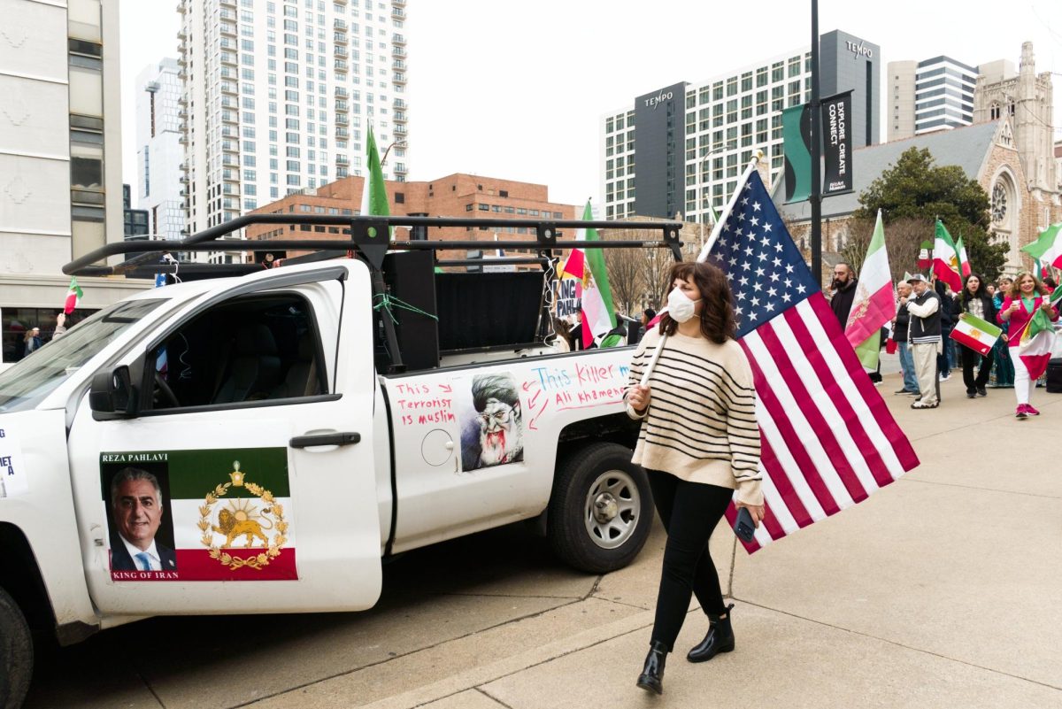 Masked demonstrator holds an American flag as she walks by a decorated pickup truck supporting Reza Pahlavi, as photographed Feb. 14, 2026. (Hustler Multimedia/George Albu)