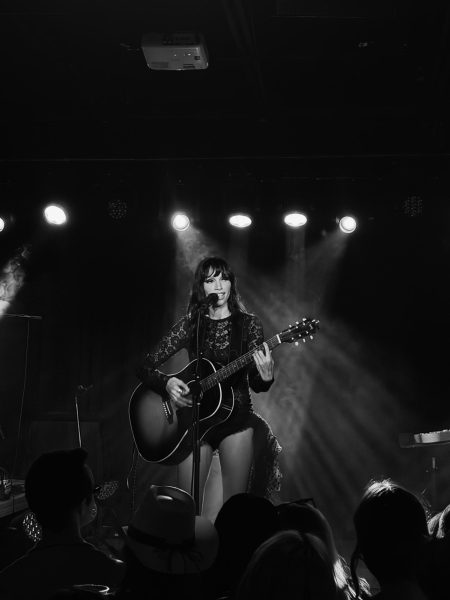 Syd Taylor holds her guitar close as stage lights cut through the haze behind her, as photographed Feb. 27, 2026. (Hustler Multimedia/Naomi Wang)